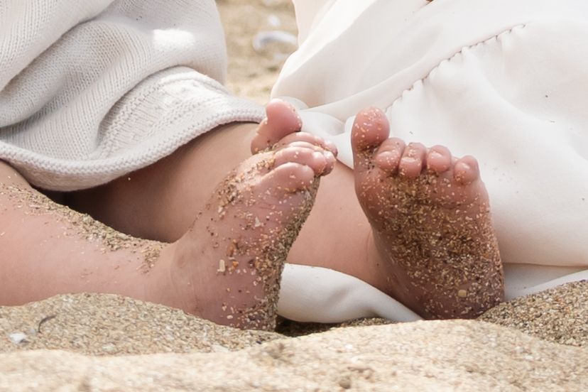 Small baby feet covered in sand and white fabric on a sunny auckland beach during a warm summer day.