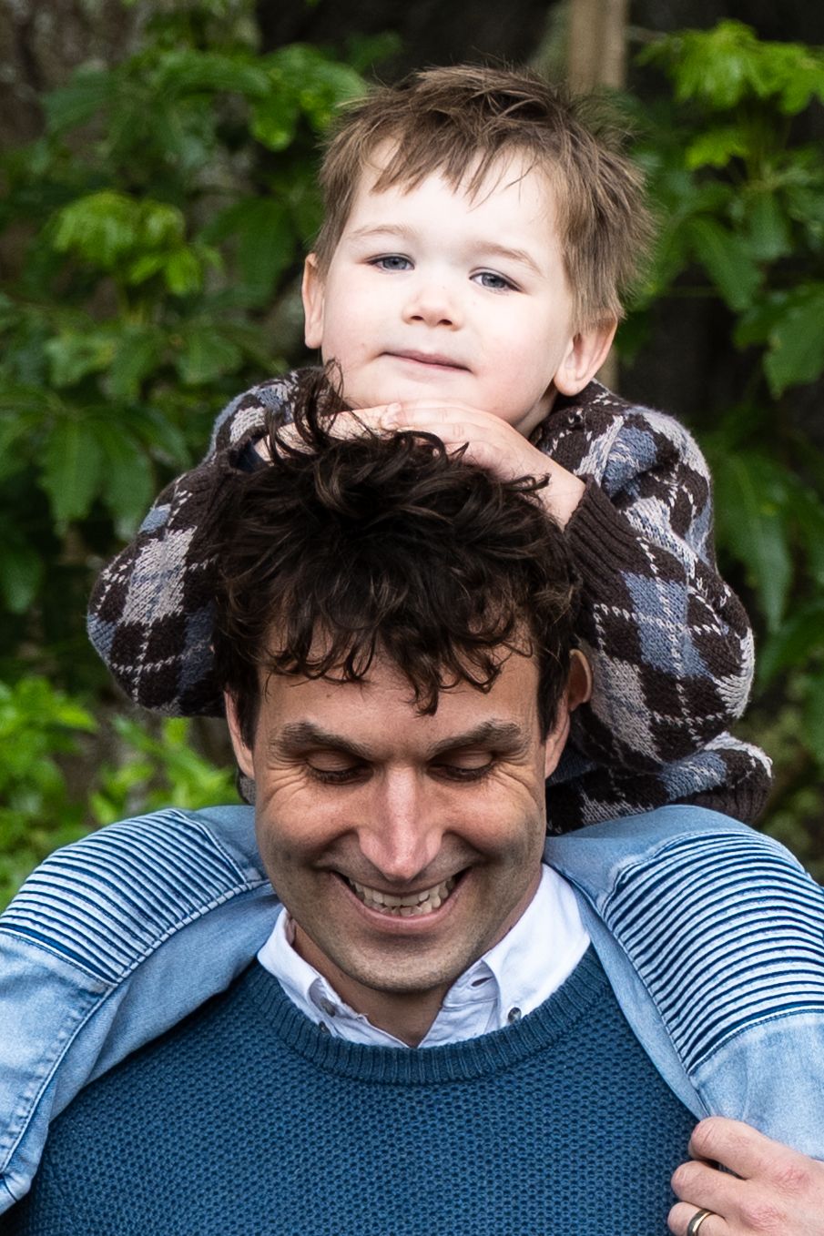 A joyful outdoor family moment shows a shoulder ride in front of green foliage on a sunny day.