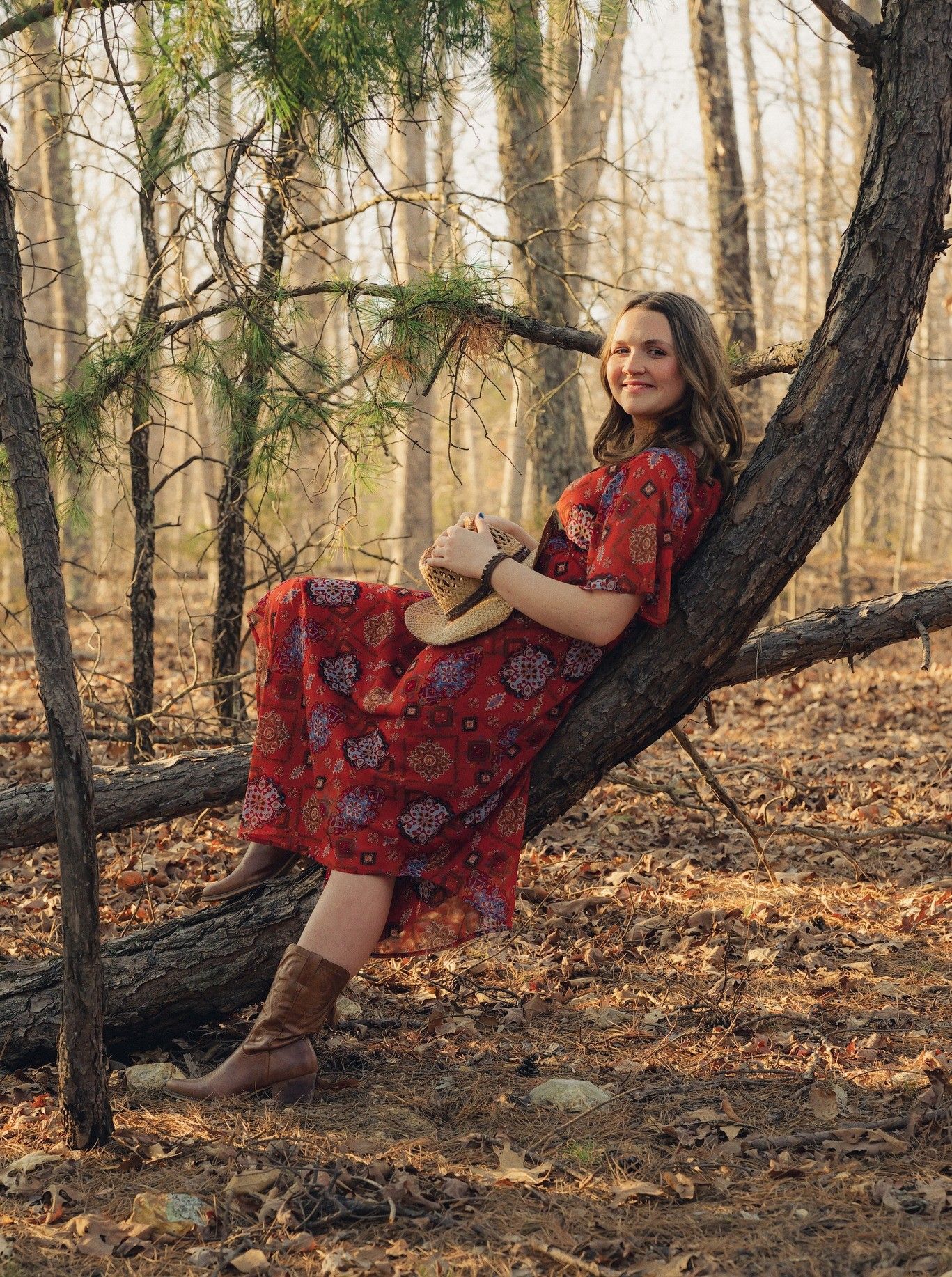 A person in a red dress reclines against fallen trees in an autumn forest setting during golden hour.