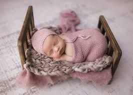 Newborn baby sleeping peacefully in rustic wooden bed wearing soft pink knitted wrap during professional photoshoot.