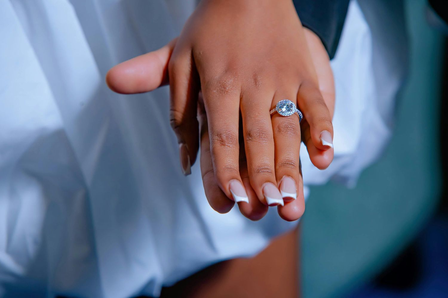 A hand wearing a sparkling engagement ring with white French manicured nails against a light background.