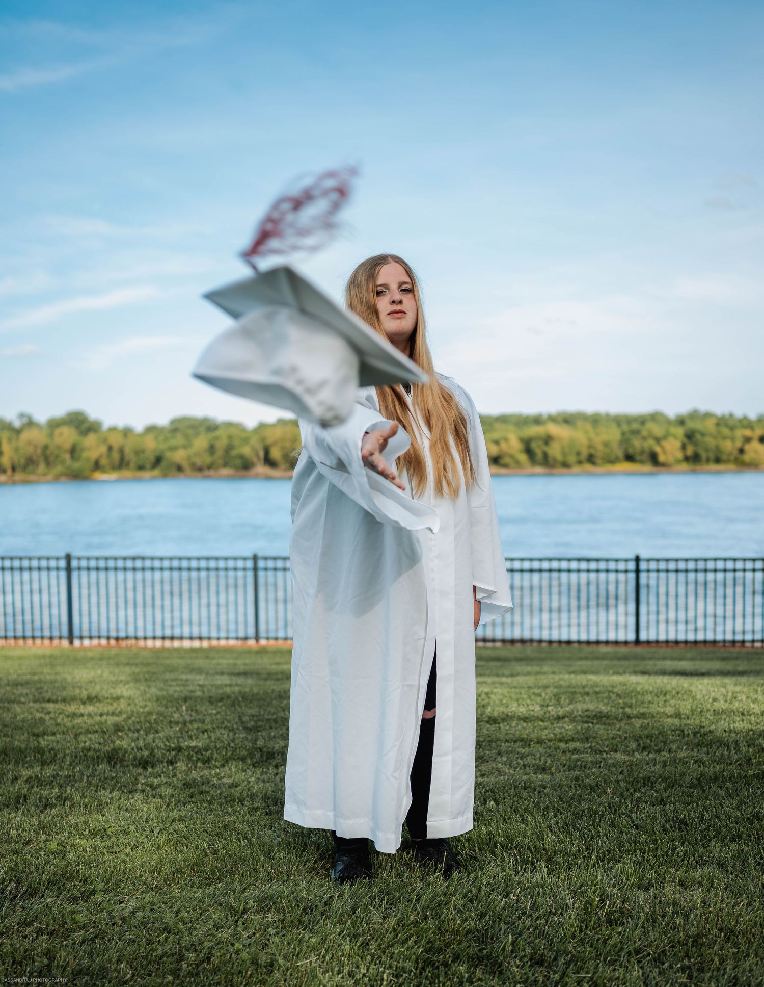 A person in a white coat twirls joyfully on green grass near a lakefront on a bright sunny day.