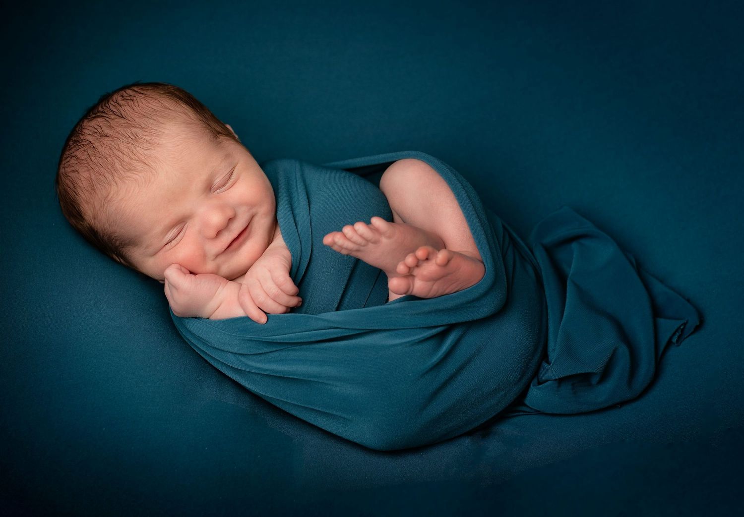 Newborn baby sleeping peacefully wrapped in dark teal blanket against deep blue background.