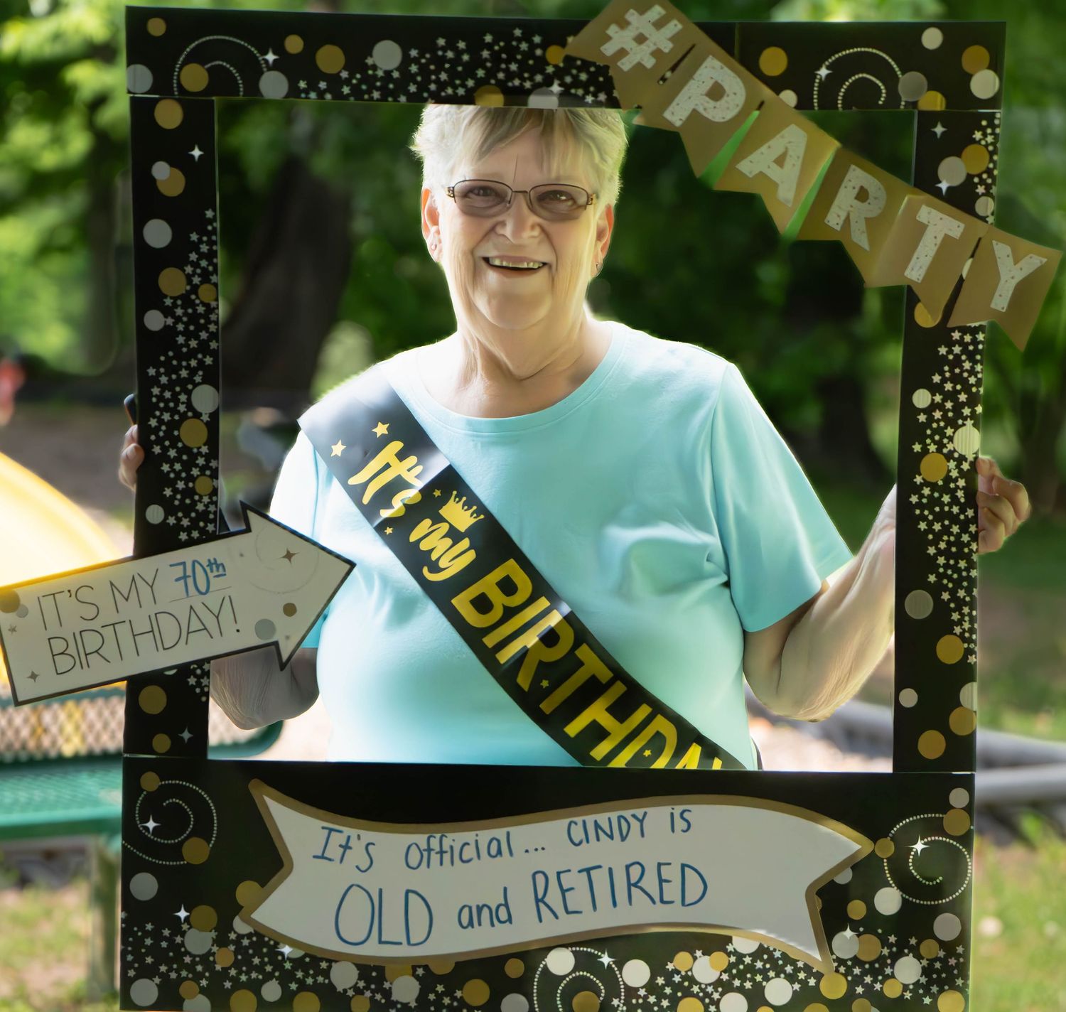 Person wearing a turquoise shirt and birthday sash celebrates retirement with decorative party banners outdoors.