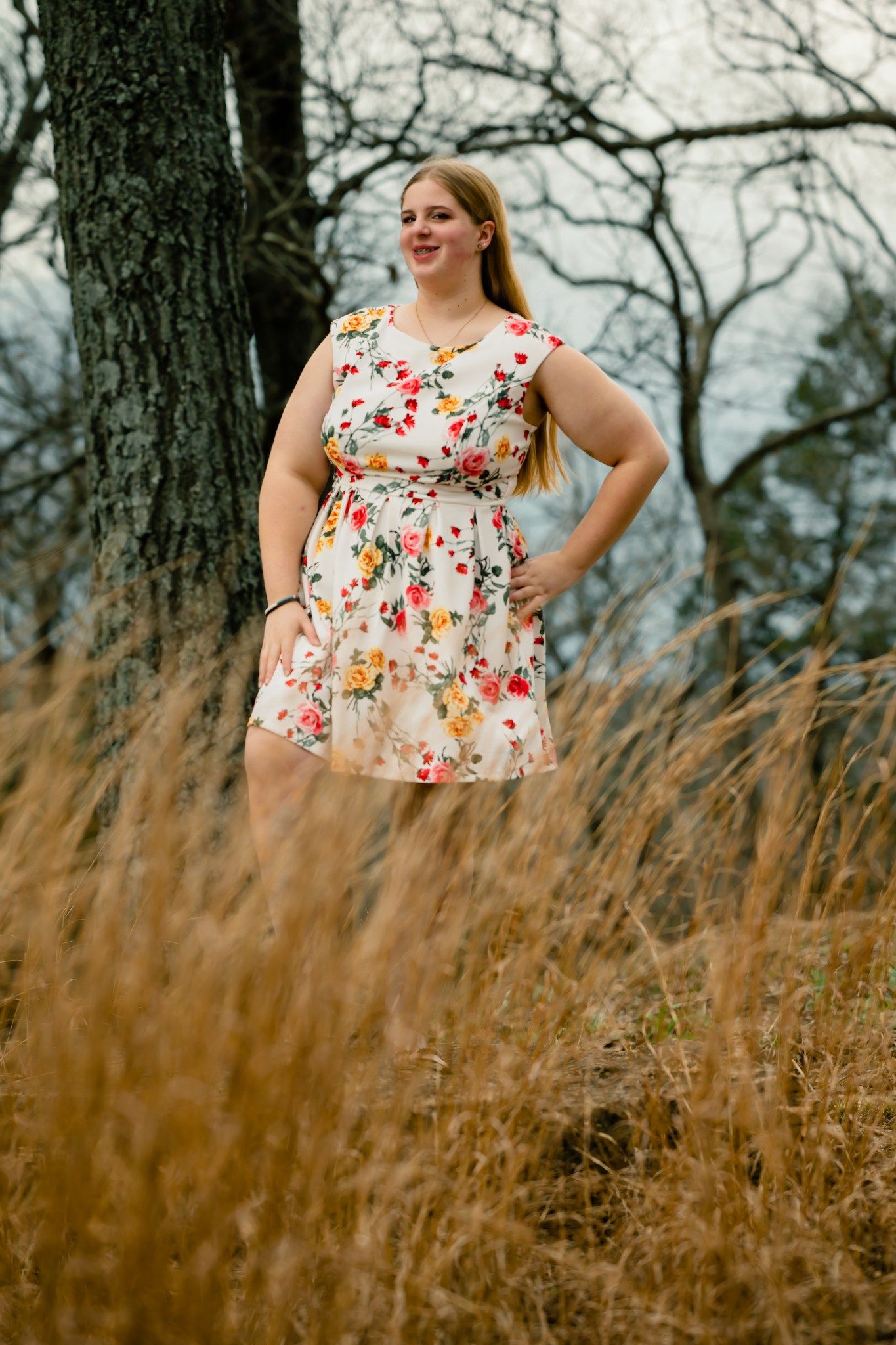 Person in a floral print sleeveless dress poses in a field of tall brown grass with bare trees in the background.