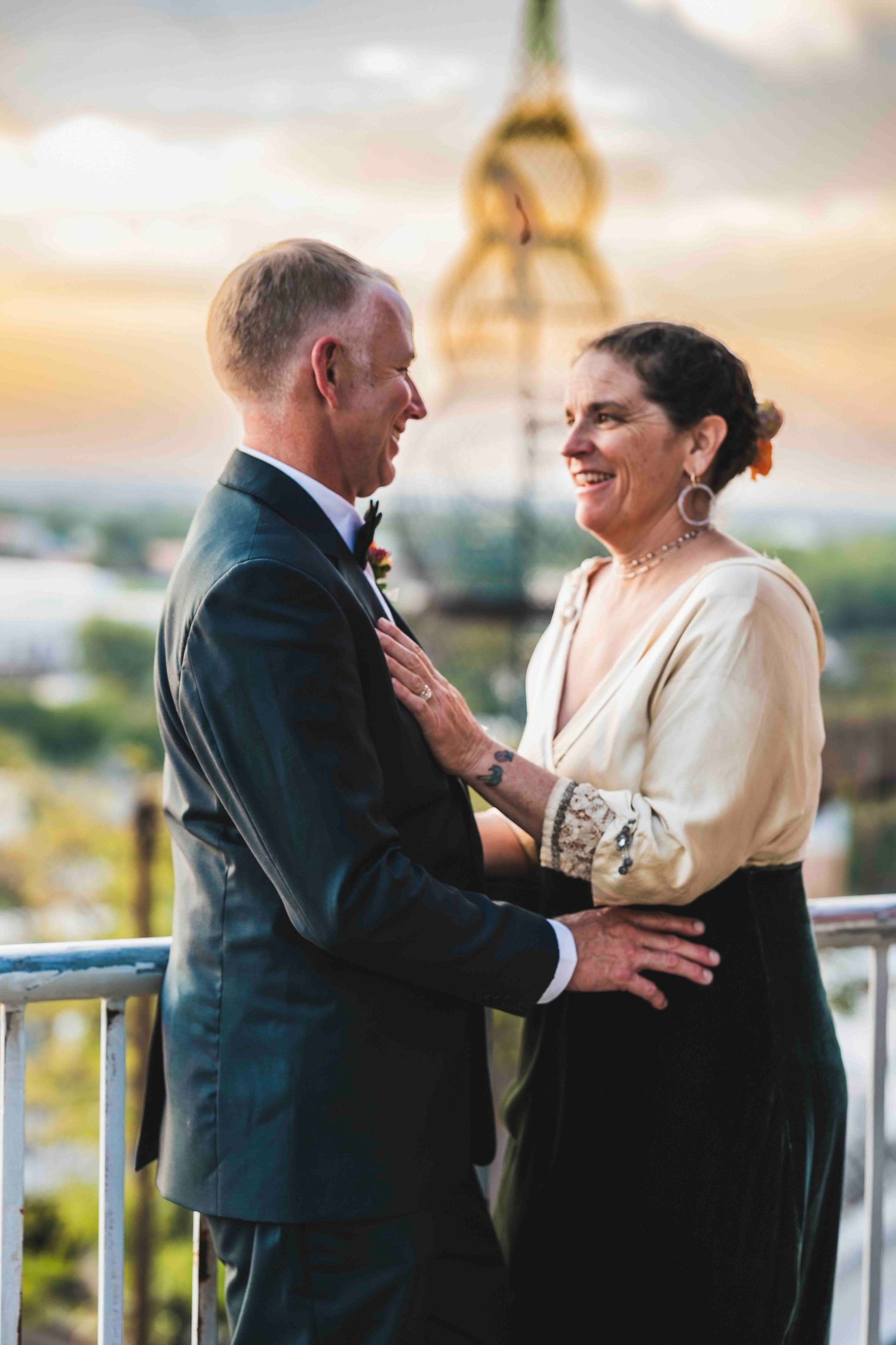 A couple shares a tender moment on a balcony during sunset, with the man in a suit and woman in formal attire.