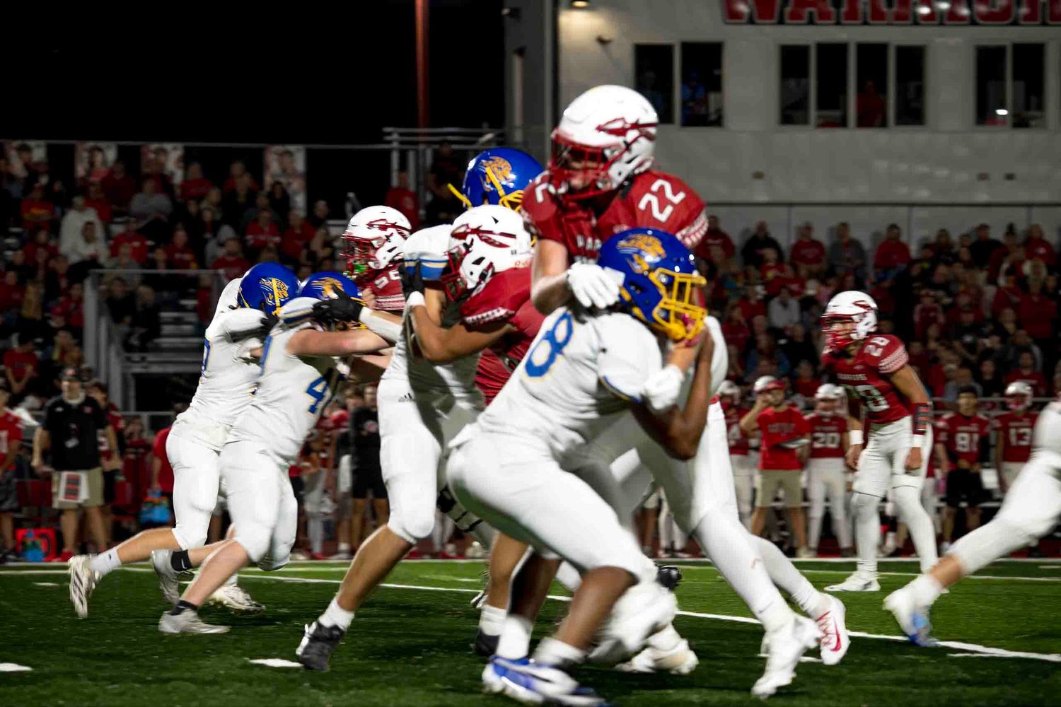 College football players engage in a high-intensity play during a nighttime game at a packed stadium.