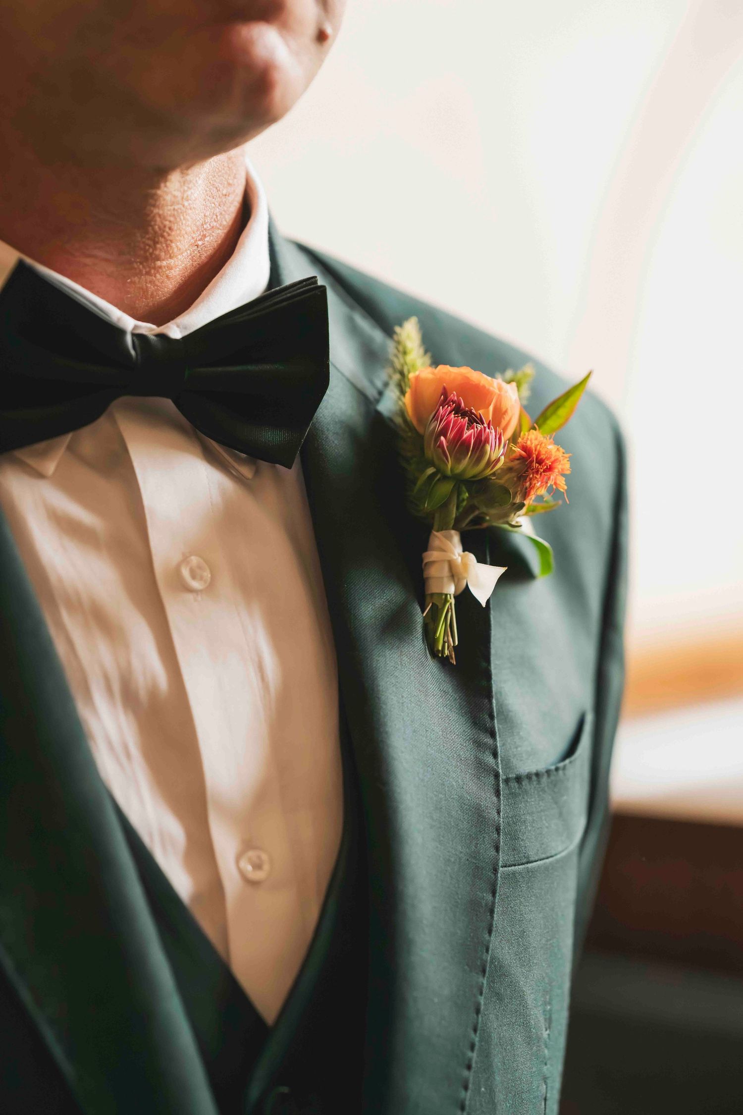 Close-up view of orange and pink floral boutonnieres on dark green suit jackets with white shirts and black bow ties.