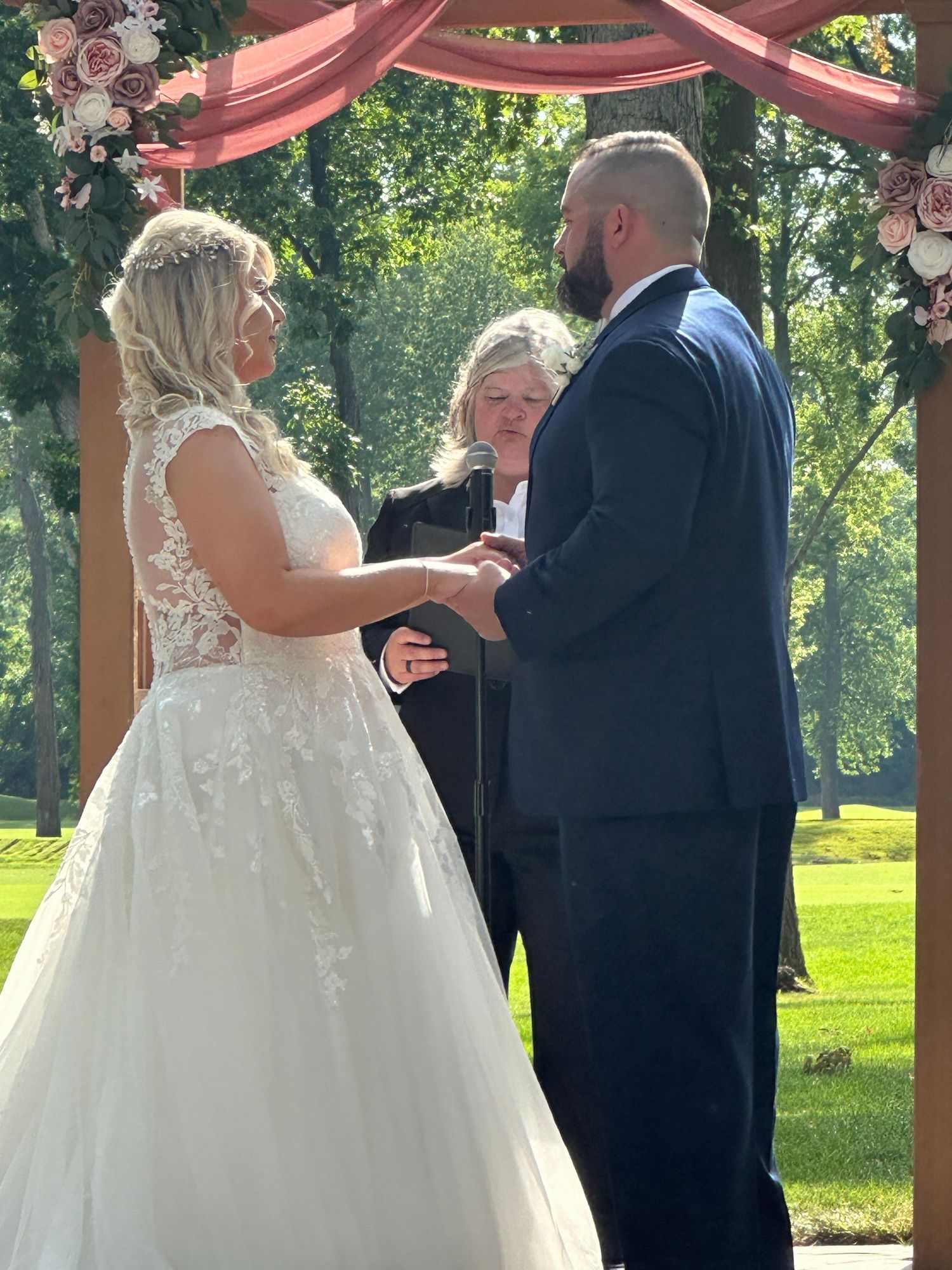 A wedding ceremony takes place under a coral-colored arch decorated with flowers in an outdoor garden setting.