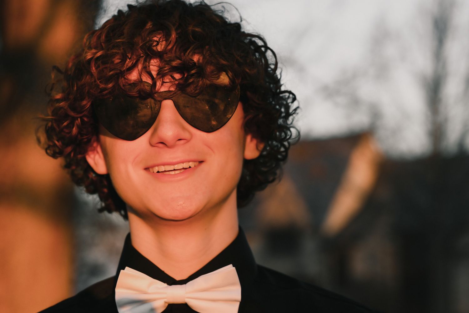 A person with curly dark hair and sunglasses smiles while wearing a black bow tie in a formal setting.