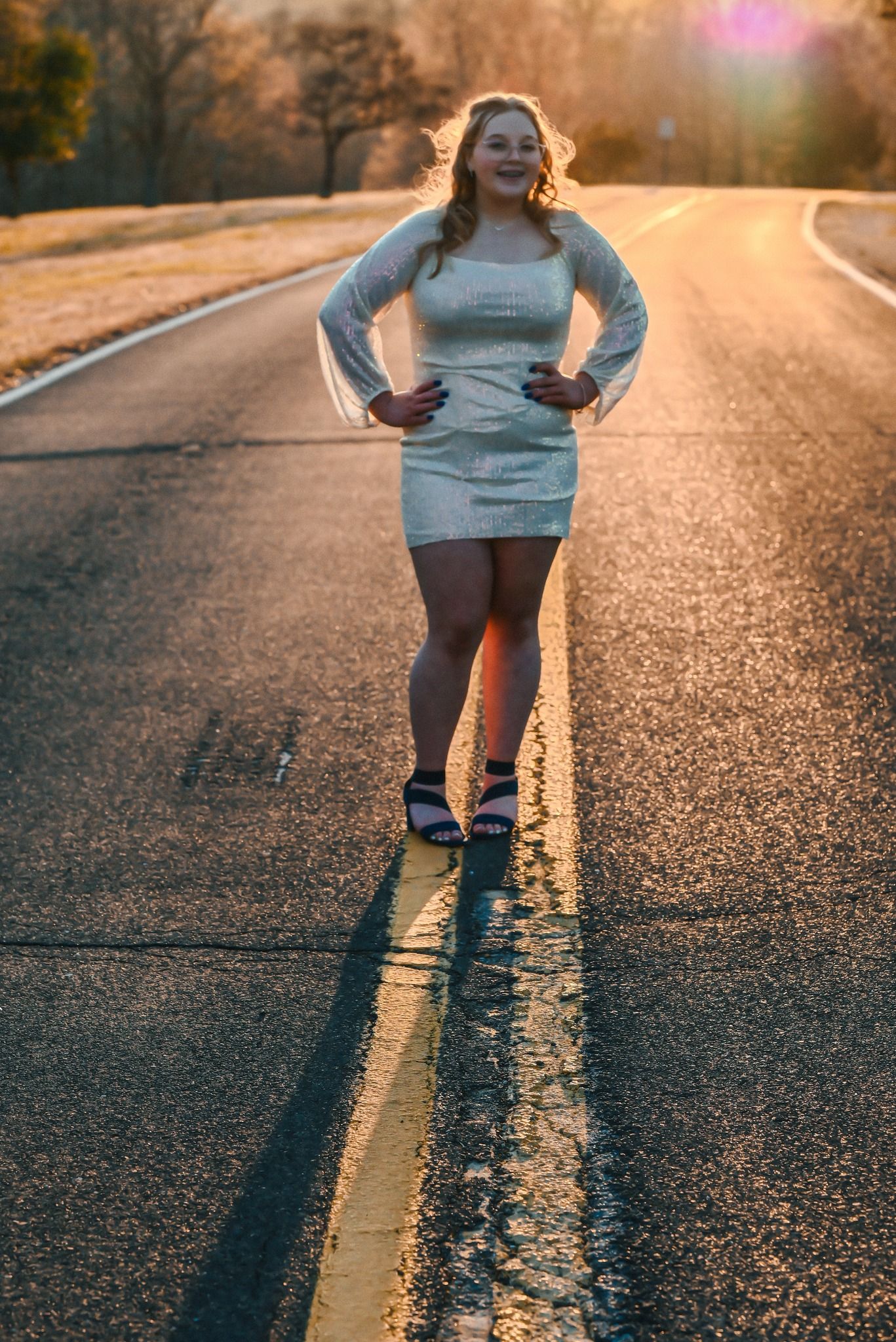 A person in a gray dress poses confidently on an empty road during golden hour sunset.