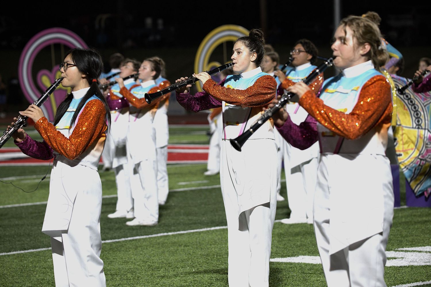 Marching band members in white uniforms perform with trumpets on a football field at night.