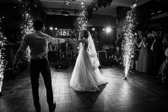 A couple shares their first dance on a dark dance floor illuminated by sparklers and stage lights at a wedding reception.