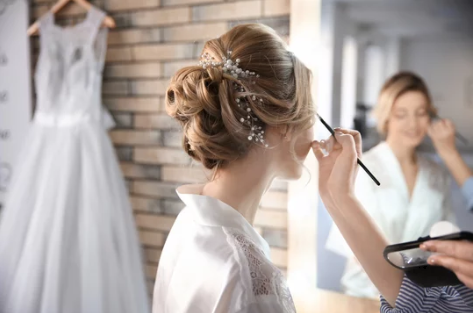 Bride getting ready with hairstylist creating an elegant updo with delicate floral accessories for her wedding day.