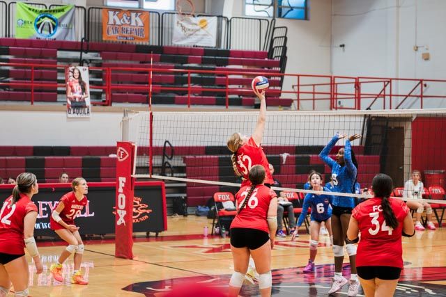 High school volleyball players compete in a match with red and blue uniforms in a gymnasium with red bleachers.