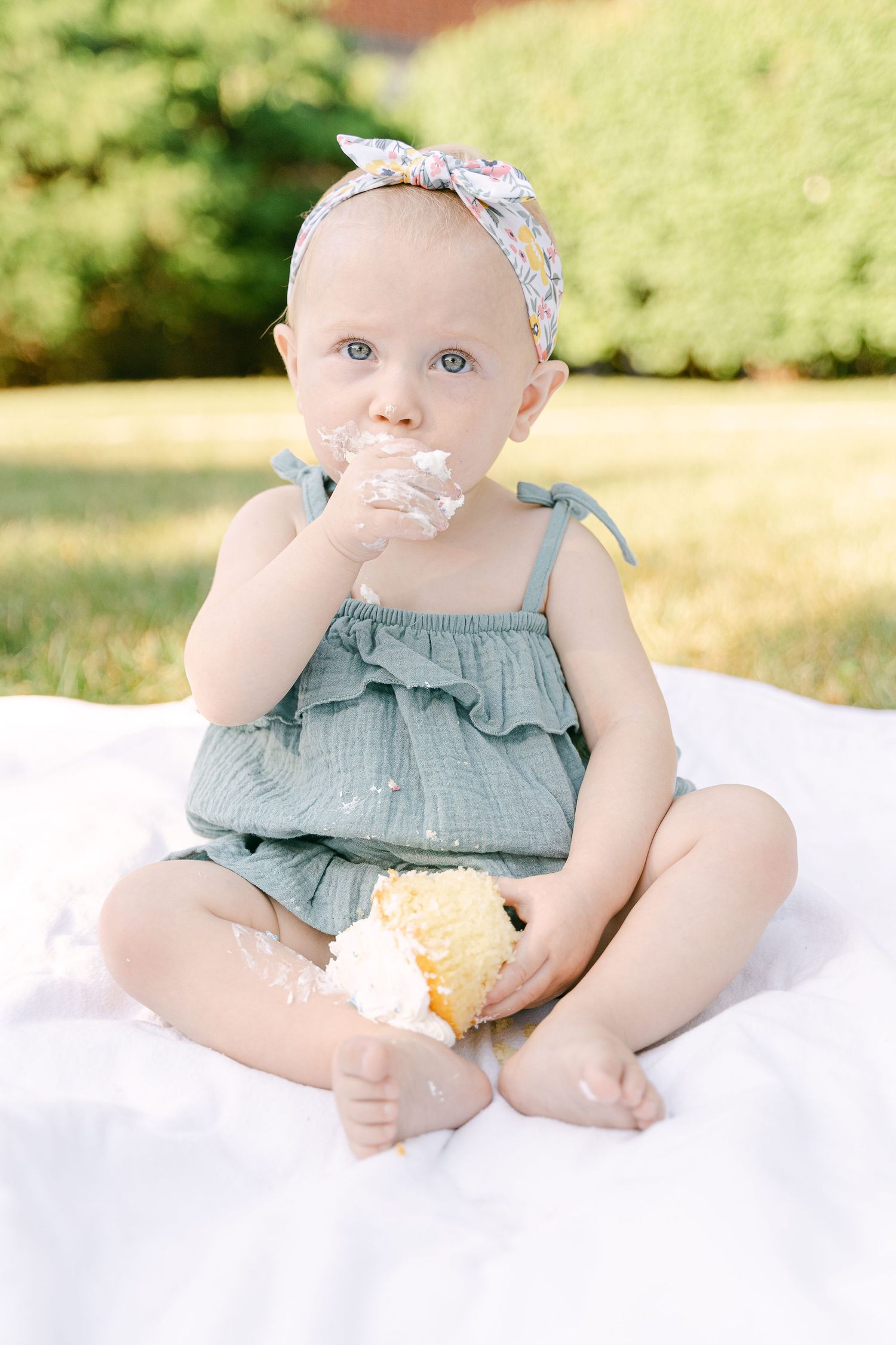 Baby in sage green outfit plays with snack while sitting on white blanket outdoors.