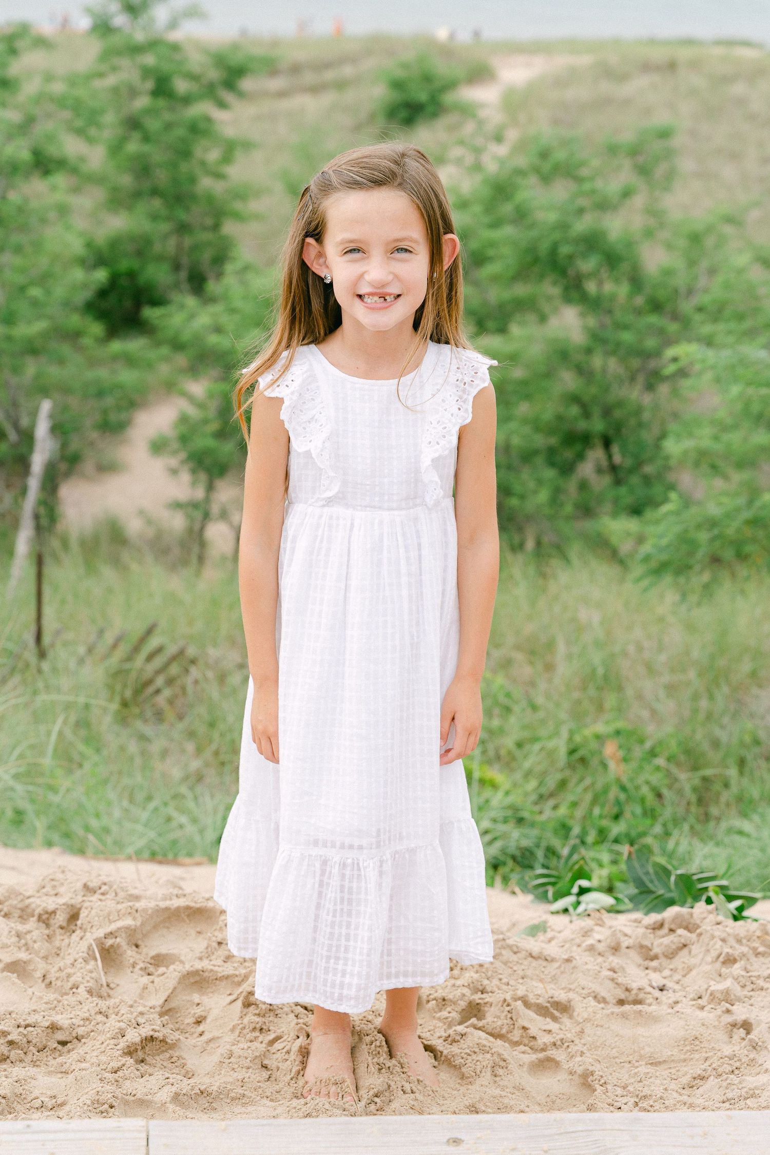 A young girl in a white summer dress stands barefoot on sandy ground with green foliage in the background.