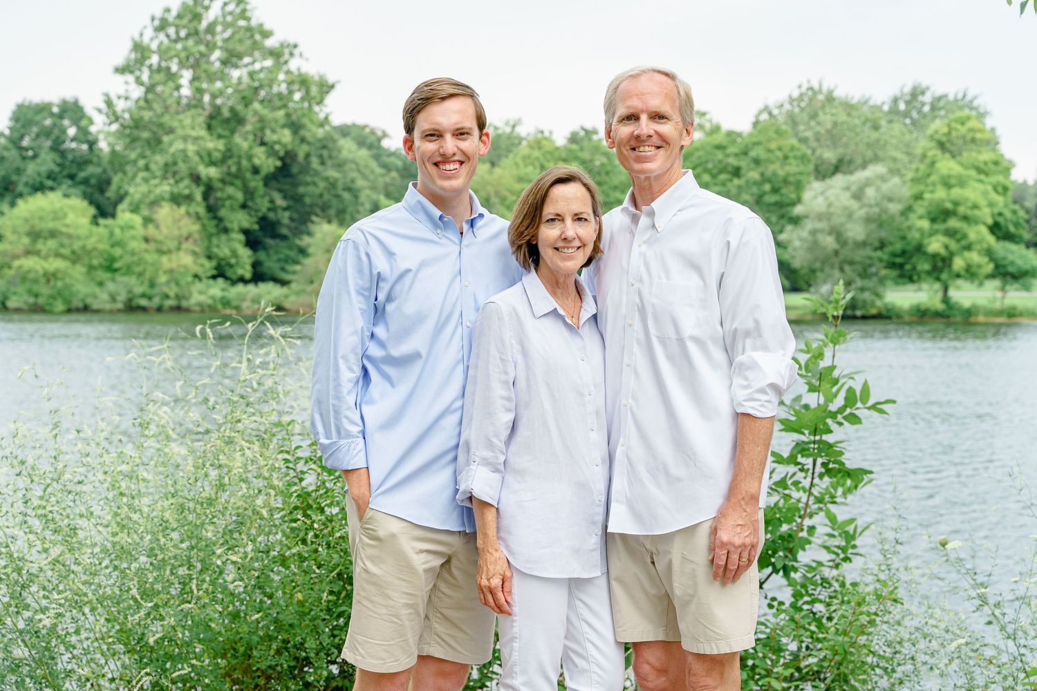 Three people stand together for a portrait wearing light colored clothing by a peaceful lake.