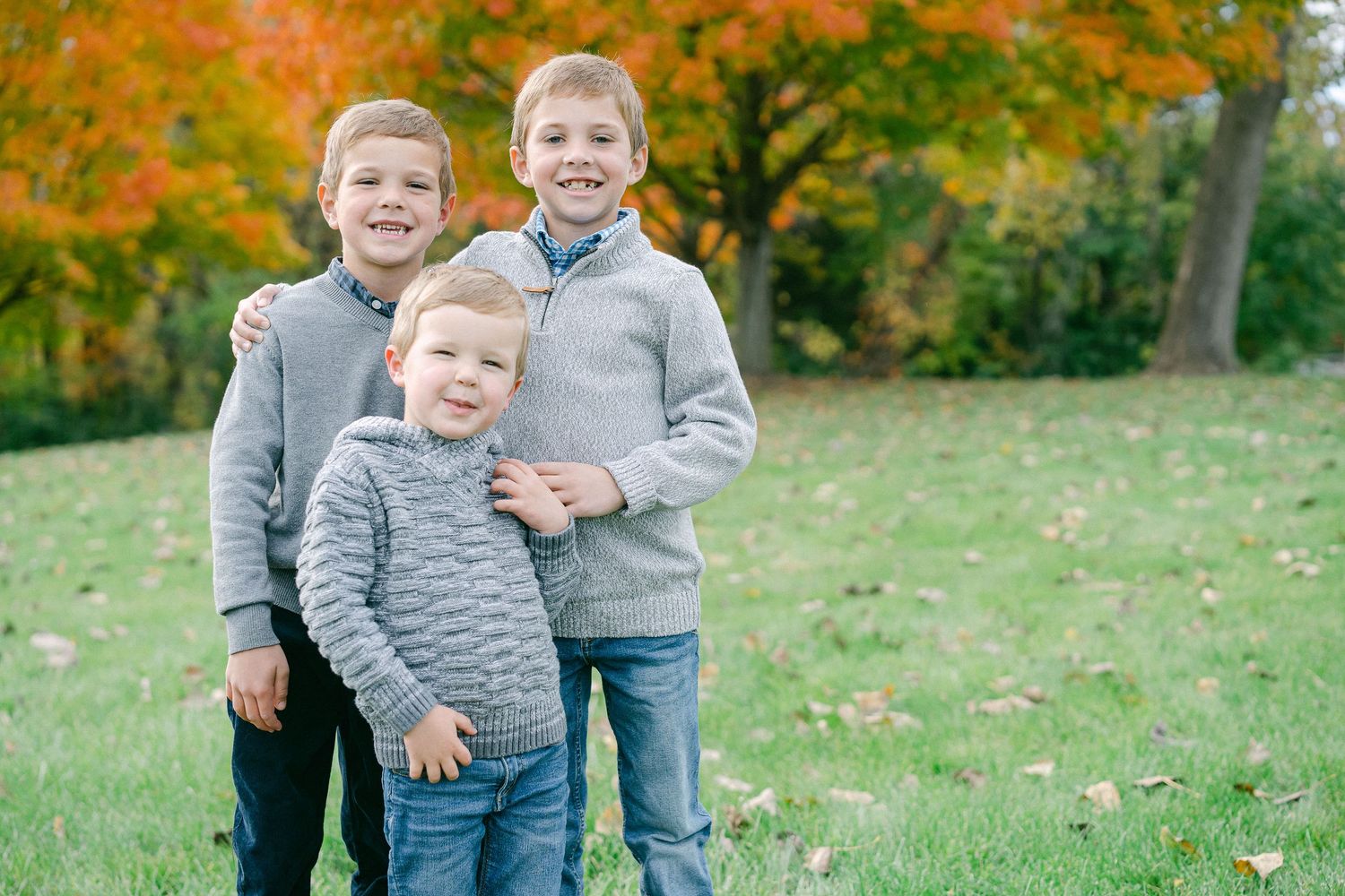 Three siblings in matching gray sweaters play together on grass surrounded by autumn trees with orange foliage.