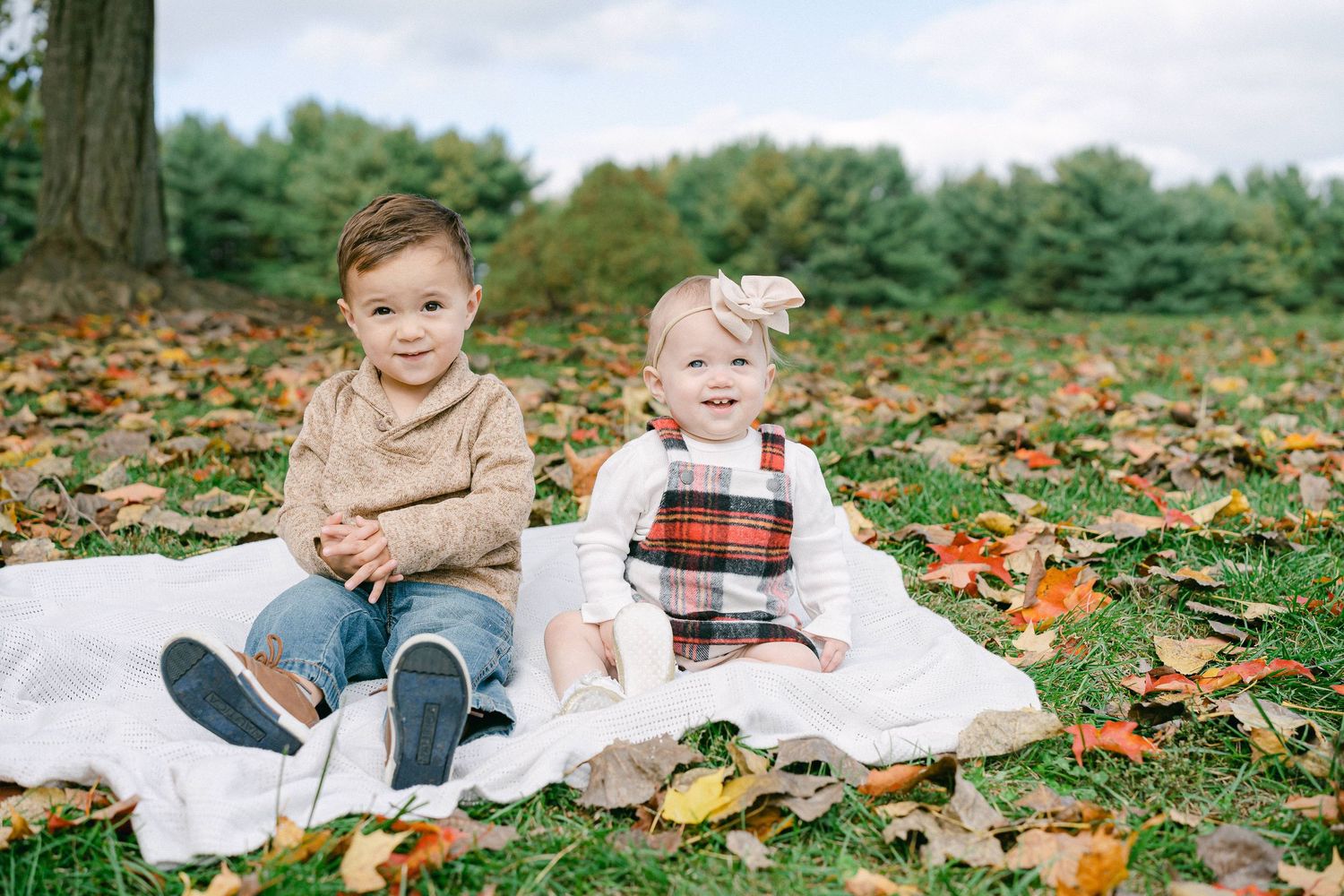 Two young children sit on a white blanket surrounded by colorful autumn leaves in an outdoor setting.
