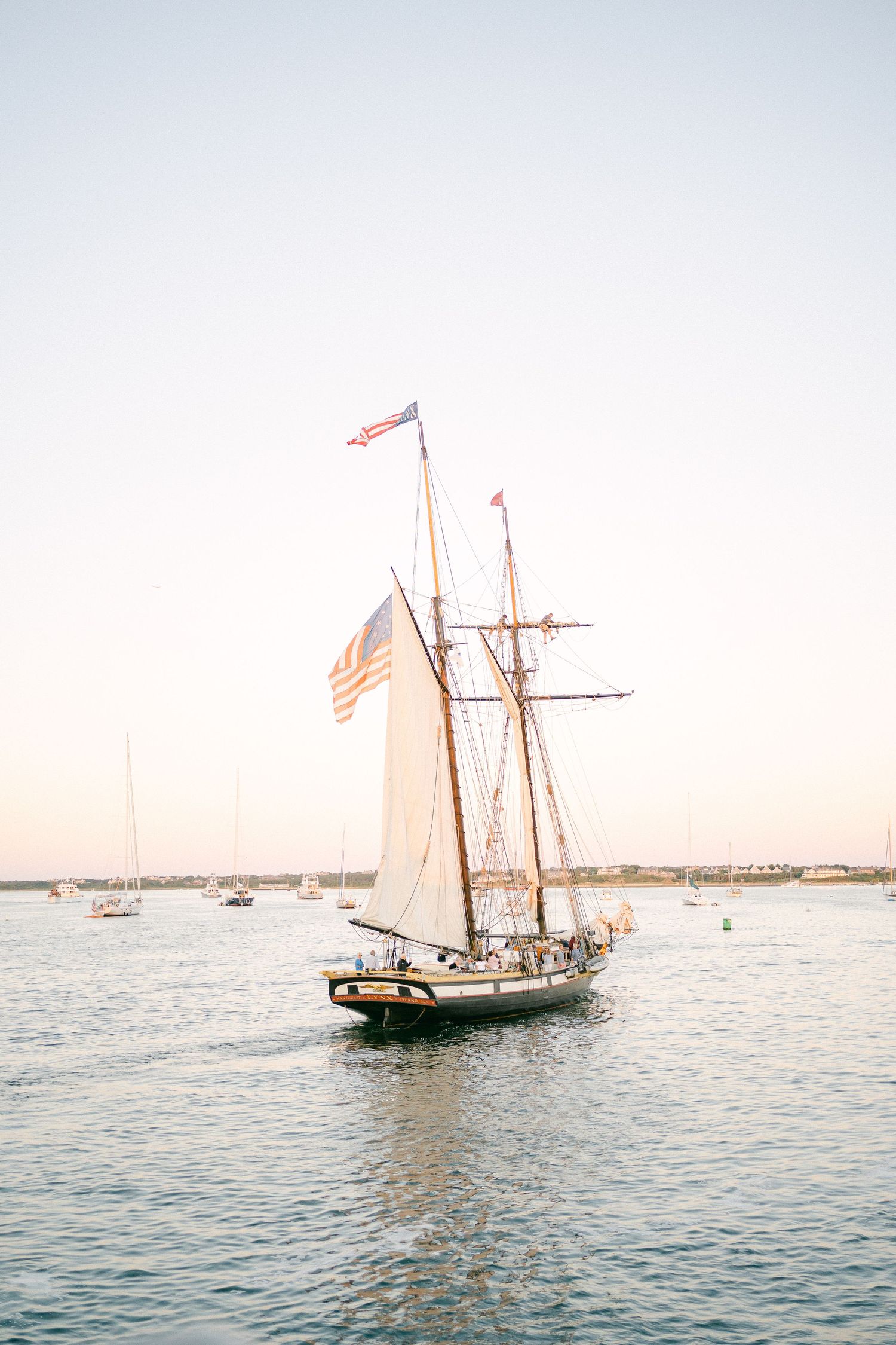 A classic sailing schooner with American flag flies through calm harbor waters at sunset.