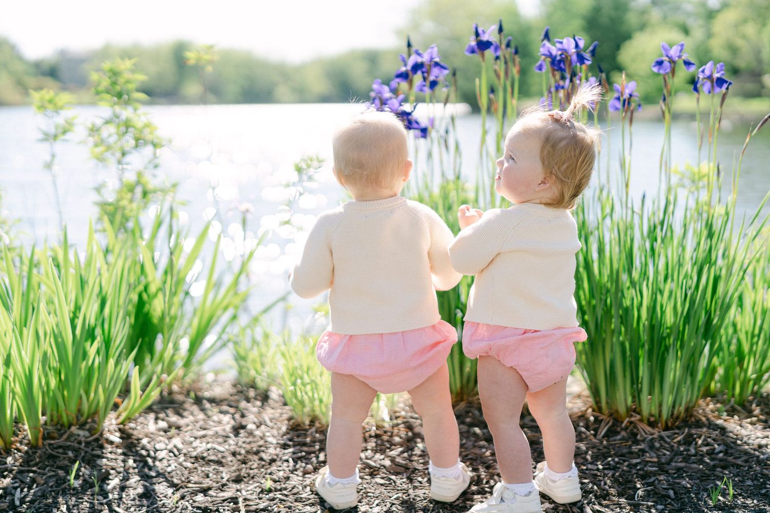 Two toddlers in pink shorts stand by a pond with purple iris flowers blooming on a sunny spring day.