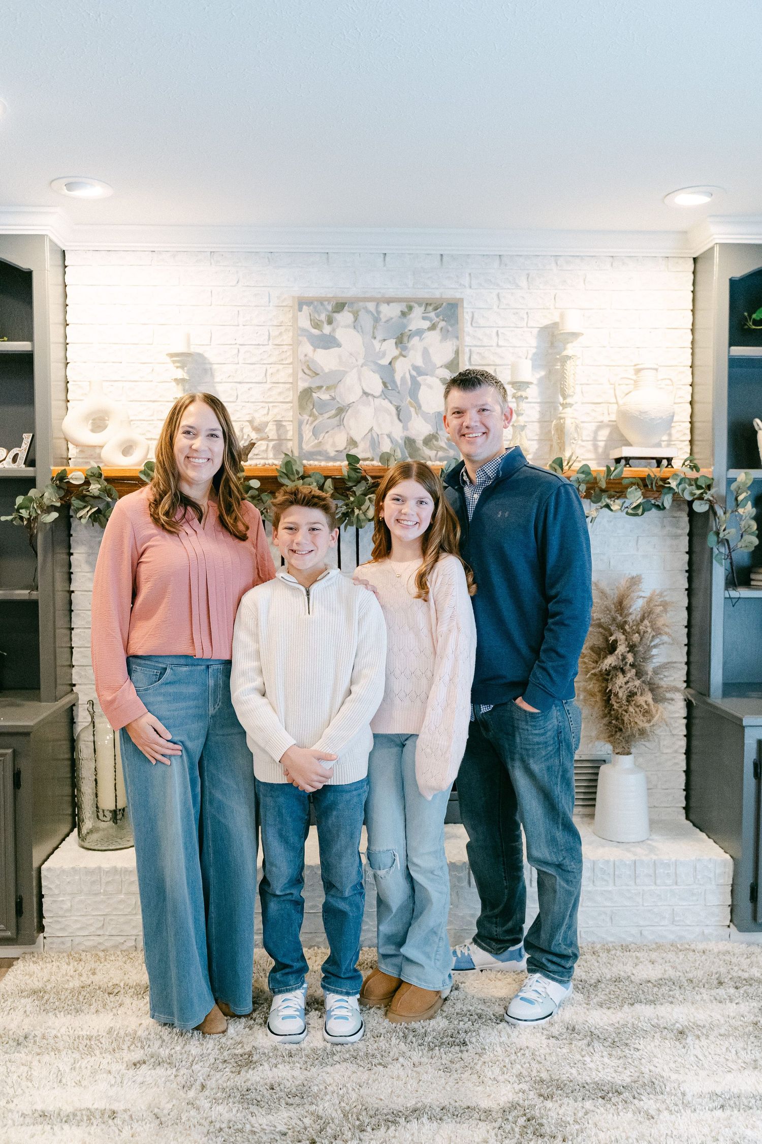 A family of four poses in front of a decorated white brick fireplace with holiday garland.