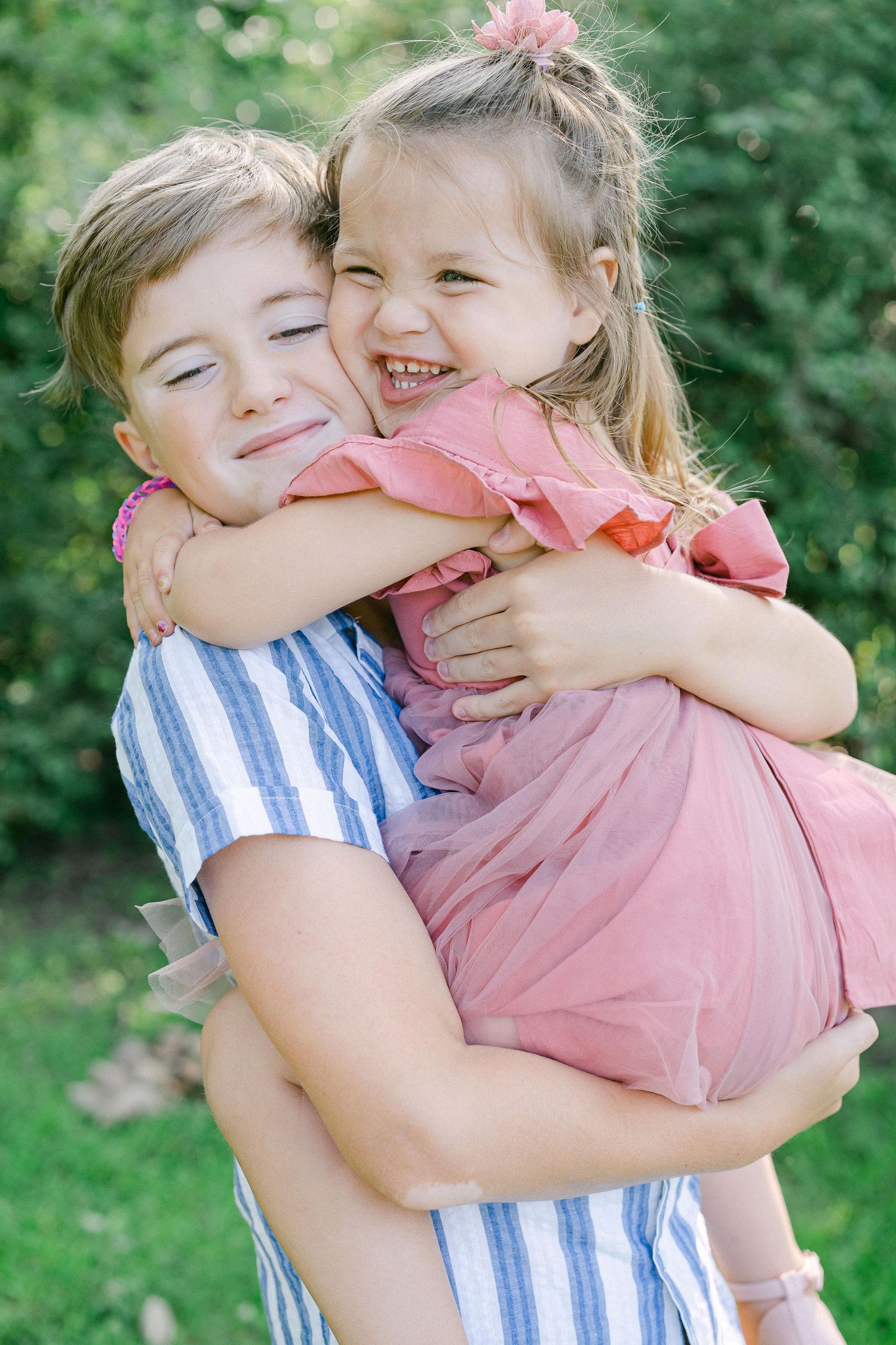 Two children share a sweet embrace while wearing pink and blue striped outfits in a sunny garden setting.