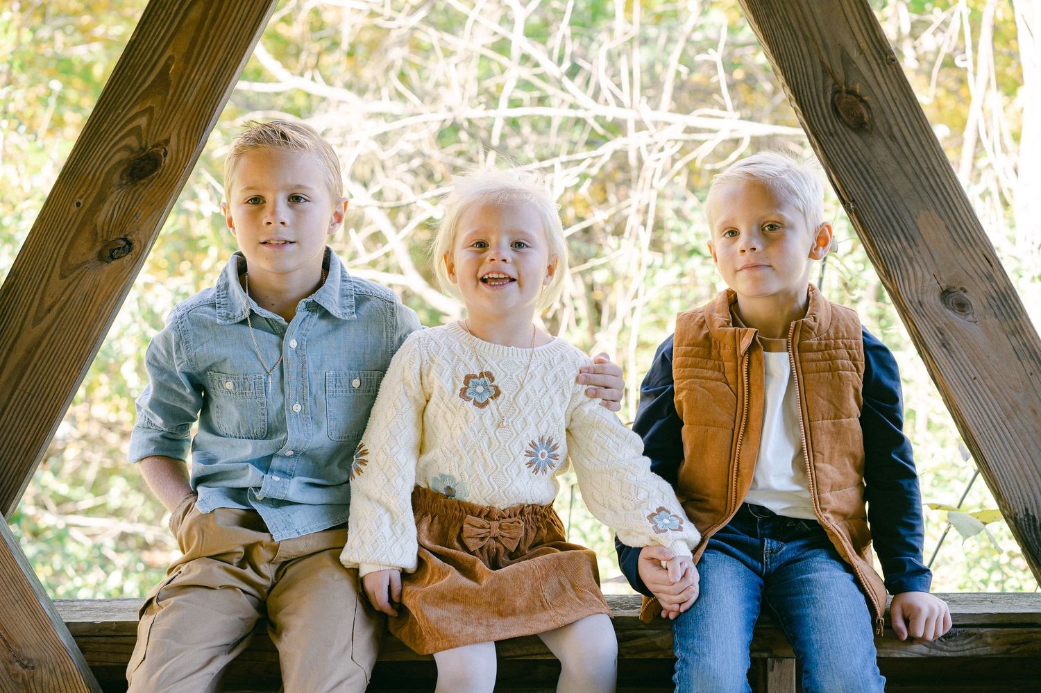 Children sit together on a wooden bench in casual clothing against a bright outdoor background.
