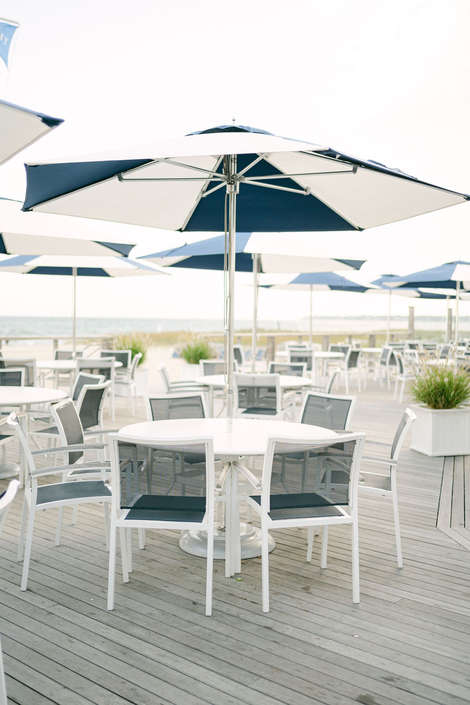 Outdoor beachside patio with white and navy blue umbrellas, round tables, and gray chairs on a wooden deck in Cape Cod