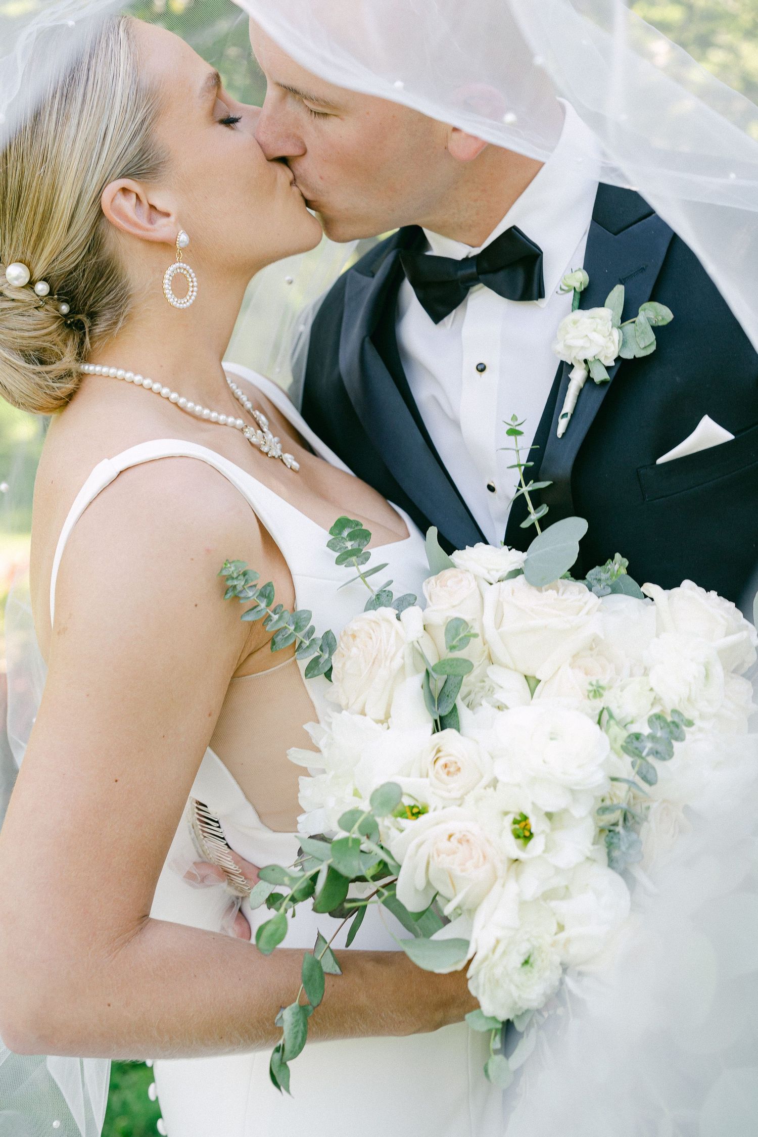 Bride and groom sharing a kiss under a veil, bride holding white floral bouquet with eucalyptus, groom in black tuxedo in Valparaiso, IN