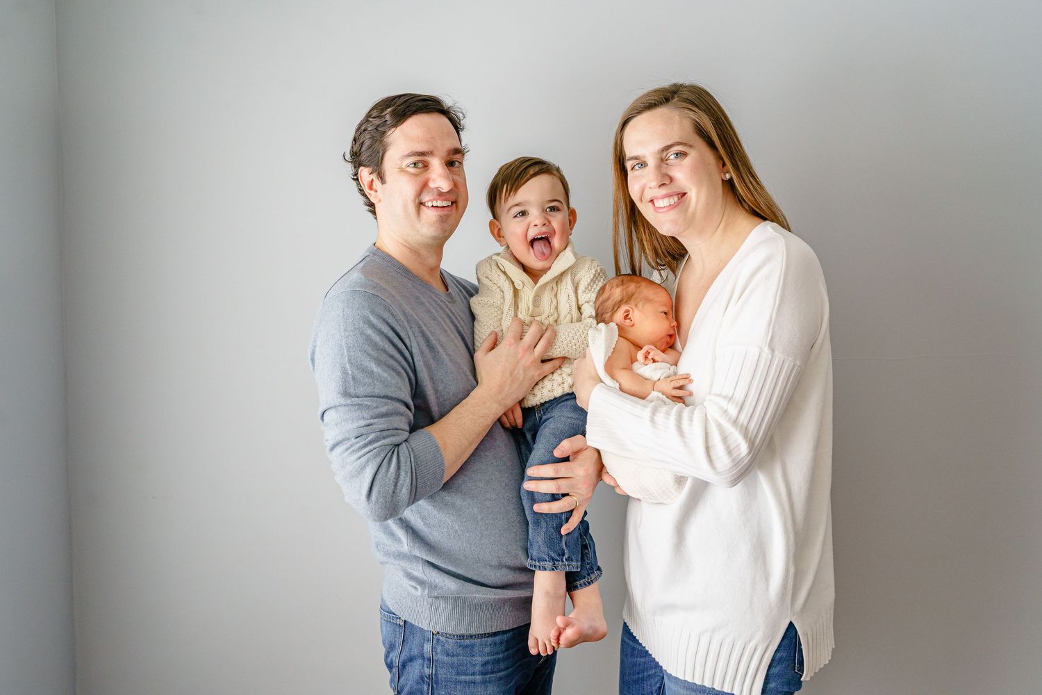 A family portrait against a white wall showing a couple with their baby and toddler.