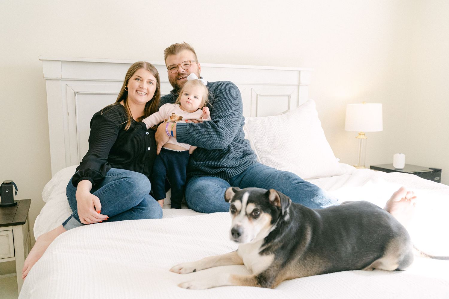 A family relaxes together on a white bed with their husky mix dog in a series of lifestyle photos.