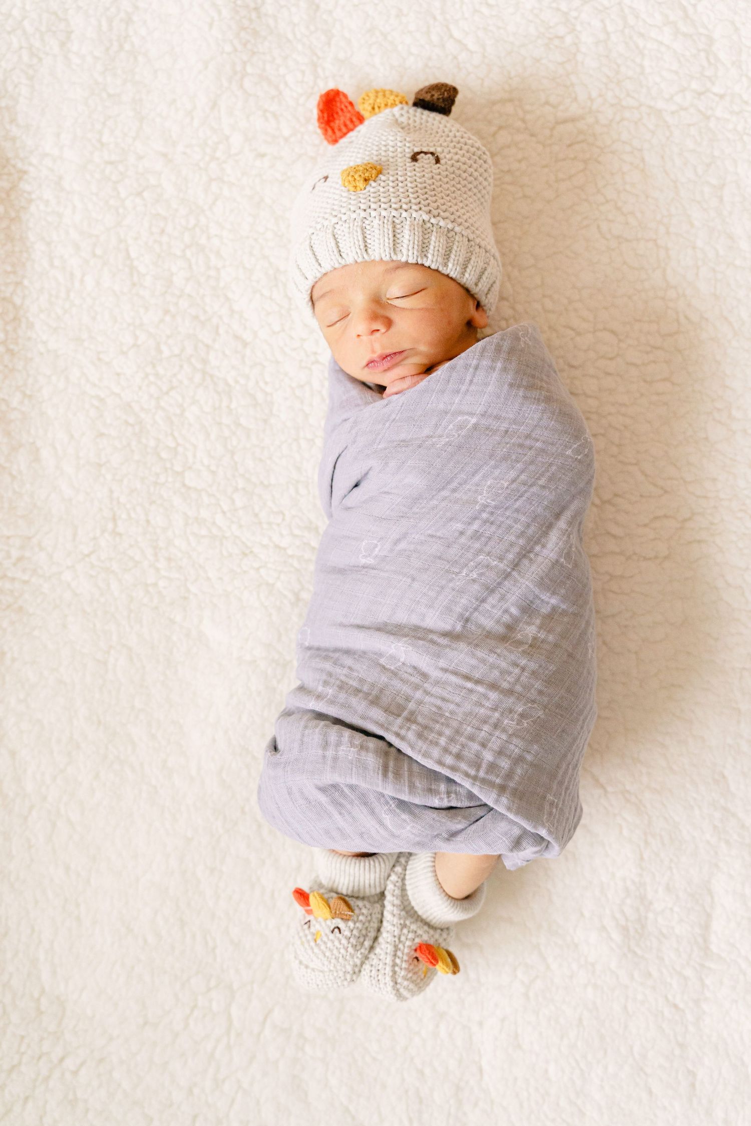 Sleeping baby wrapped in soft gray swaddle wearing cute knit animal hat and matching booties on white background.
