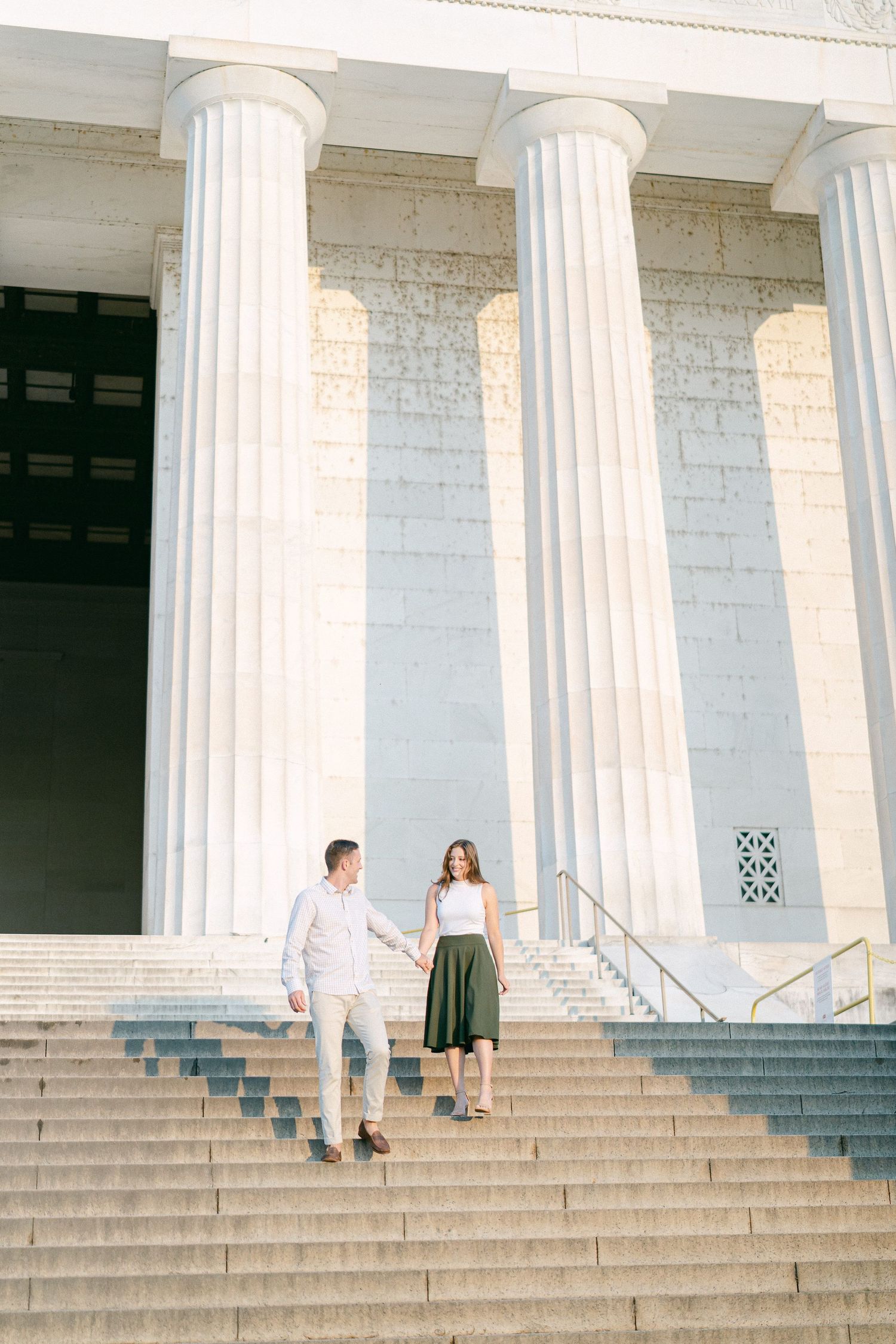A couple stands on grand white stone steps with classical columns at a historic building during an engagement photoshoot.