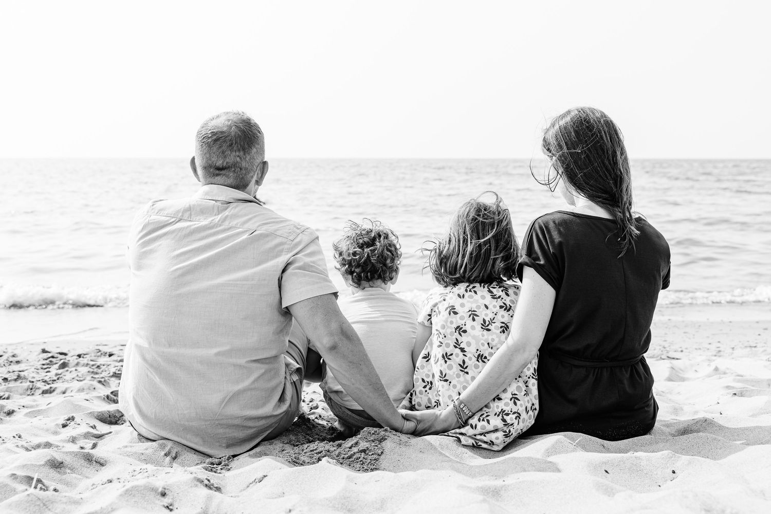 A family sits together on a sandy beach looking out at the ocean in black and white.