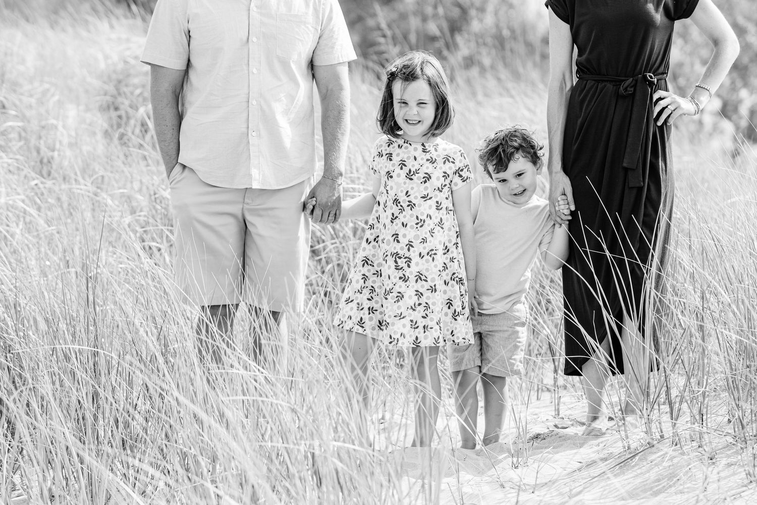 A family wearing light colored clothing poses together in a field of tall grass during a golden hour photoshoot.