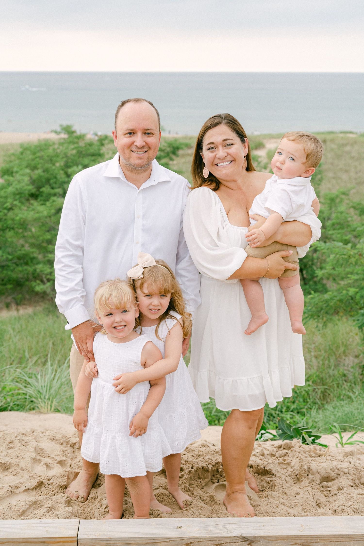 Family dressed in white poses together on sandy hill with greenery and ocean views in the background.