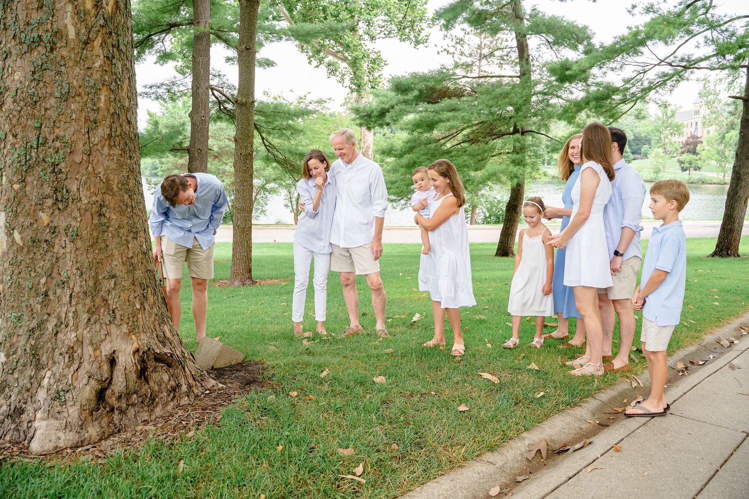 A group stands together in casual summer attire beneath tall trees in a grassy outdoor setting.