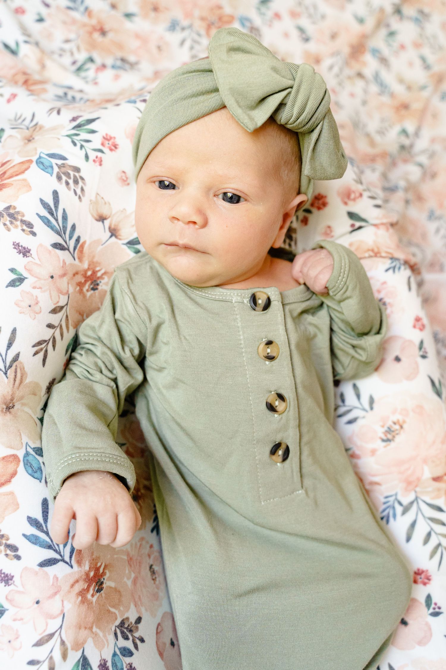 Baby wearing sage green knotted headband and matching button-up sleeper lying on floral bedding.