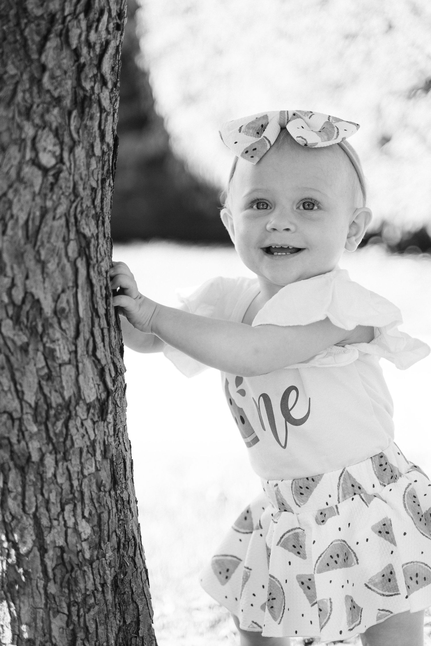 Black and white photo series showing a small child in a patterned dress playing near a tree trunk.