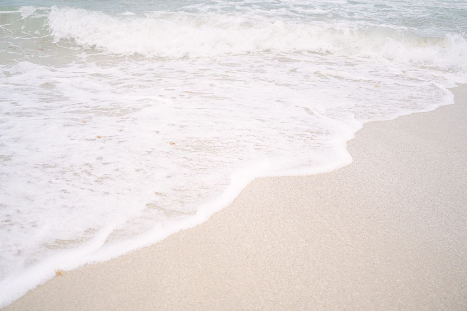 Gentle ocean waves wash up on a pristine sandy beach with white foam under an overcast sky.