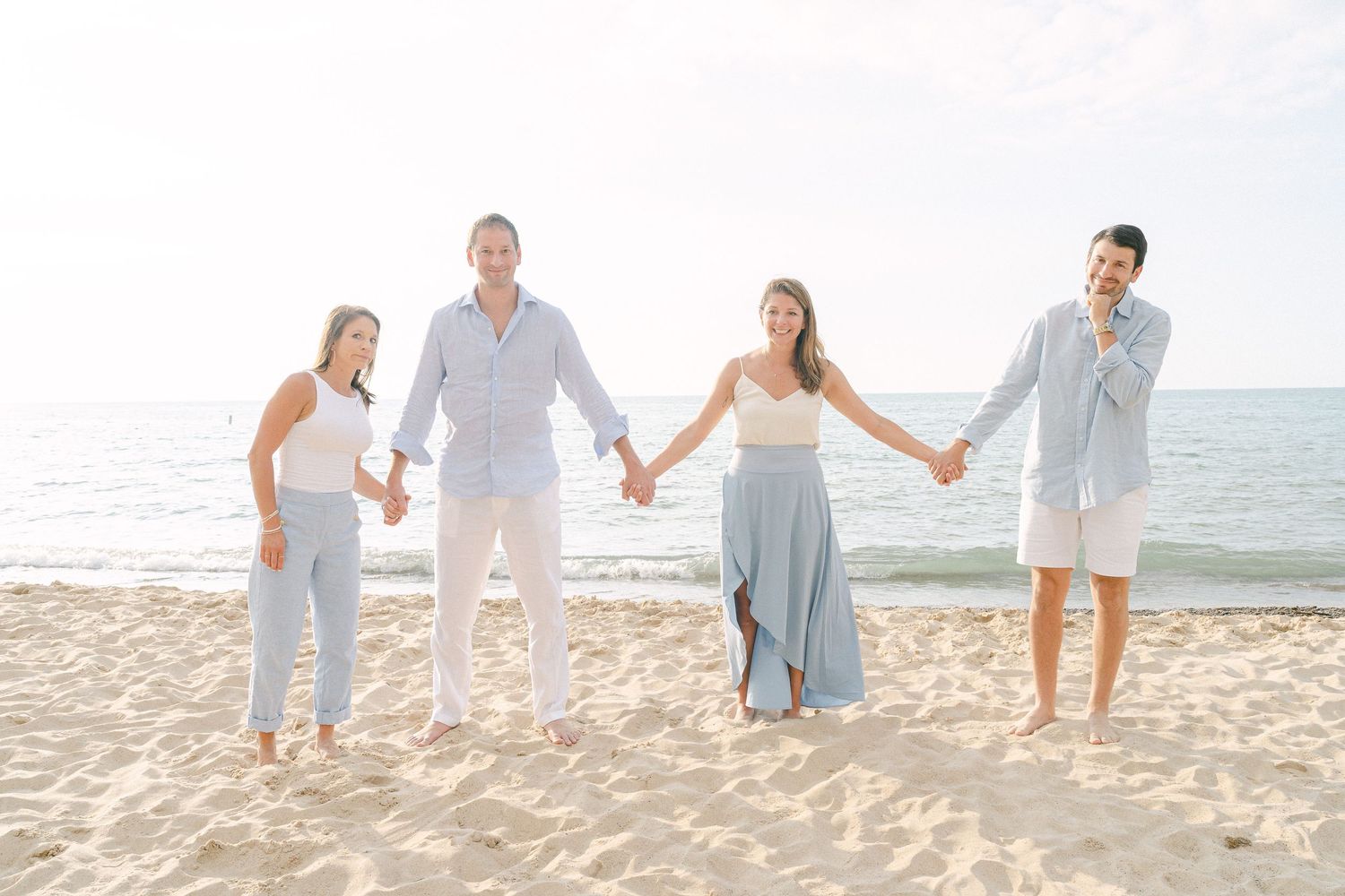 Family walking together on a sandy beach wearing light colored clothing during a sunny day by the ocean.