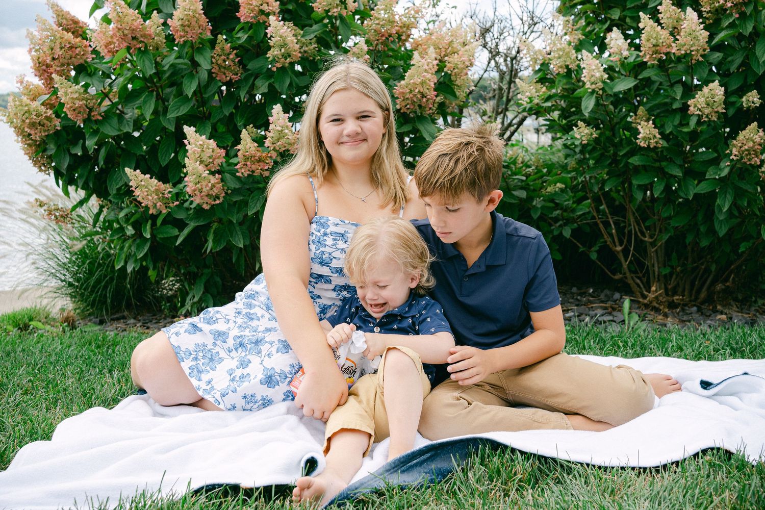 A family relaxes together on a white blanket in a lush garden setting with blooming flowers.