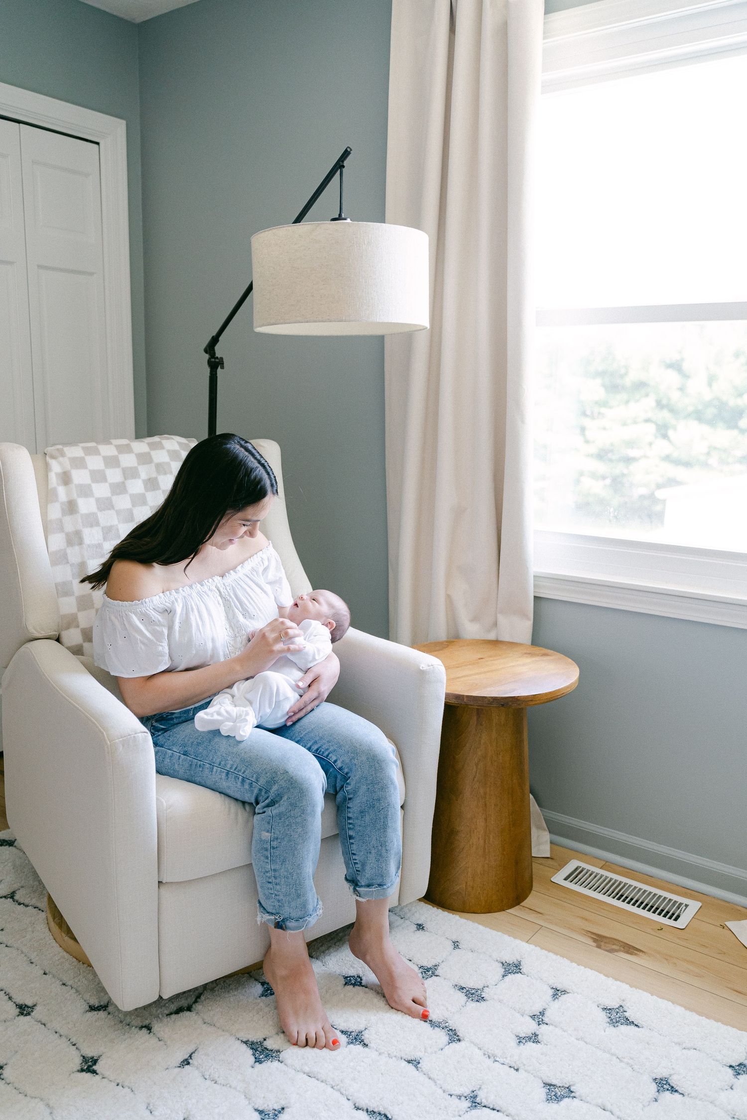 Nursery interior with white armchair, wooden side table, and blue-gray walls with white curtains.
