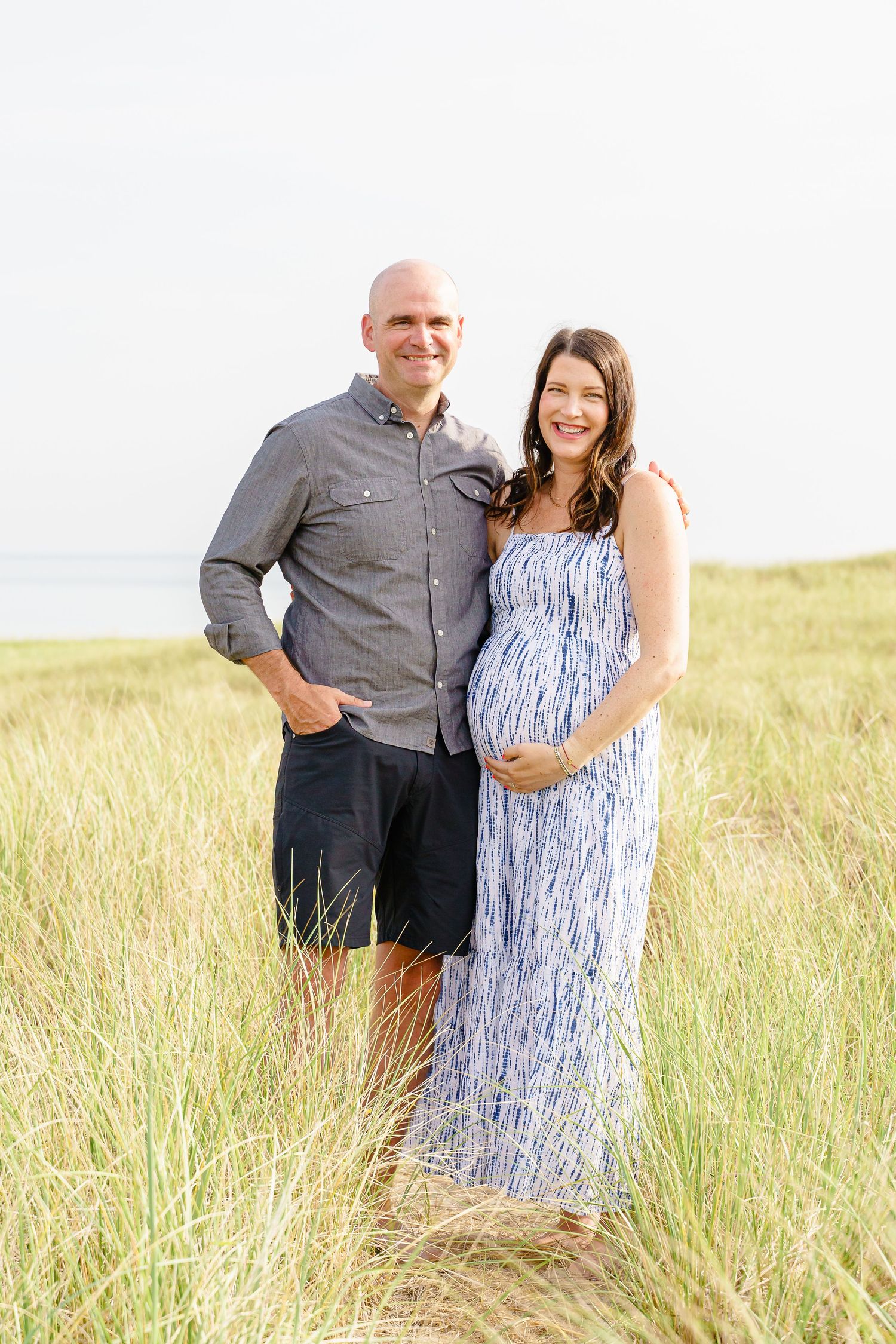 A couple stands together in a sunlit field of tall grass, wearing casual attire and embracing.