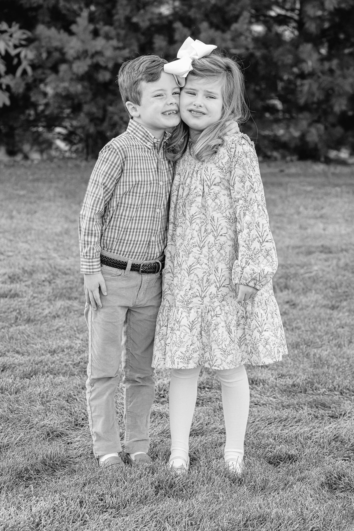 Two young children in casual attire share a sweet moment together on a grassy lawn in black and white.