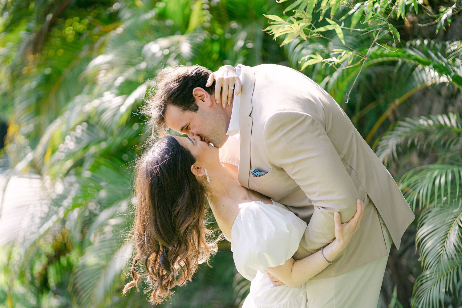 Couple sharing a romantic kiss outdoors, groom in beige suit dipping bride in white dress among lush tropical palms, in Miami, FL