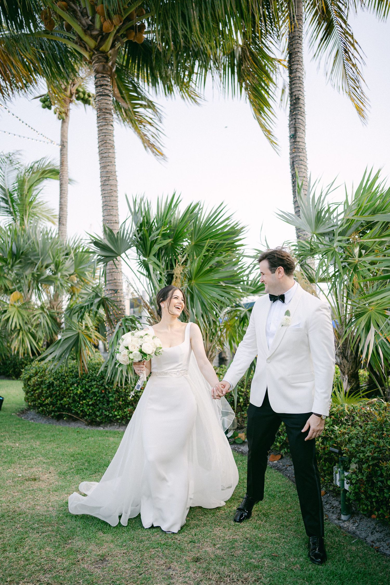 A bride and groom hold hands while walking through a tropical garden with palm trees, she in a white gown, he in a white tuxedo in Miami, FL