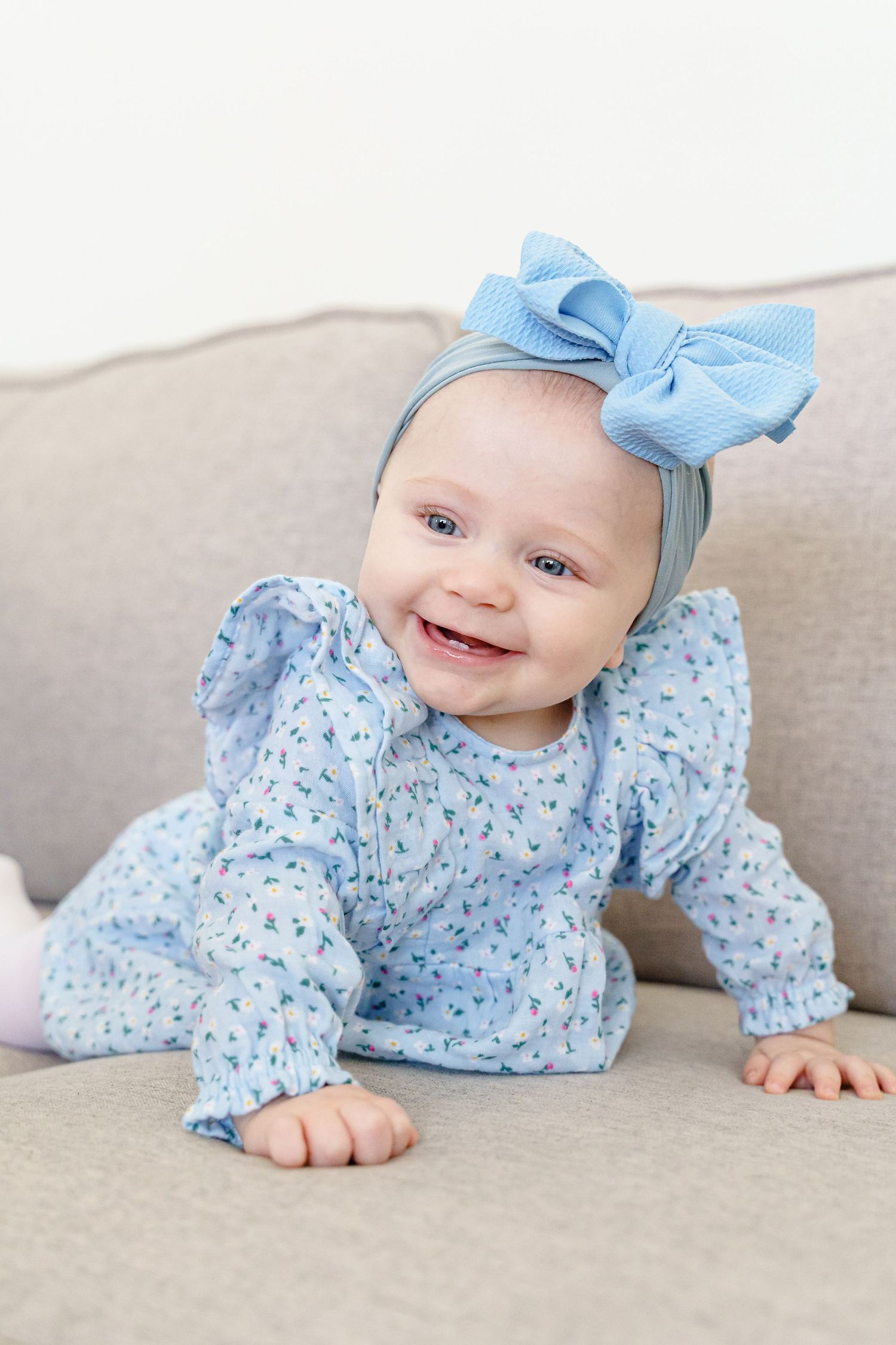 Smiling baby in blue floral outfit and matching bow relaxing on gray sofa.